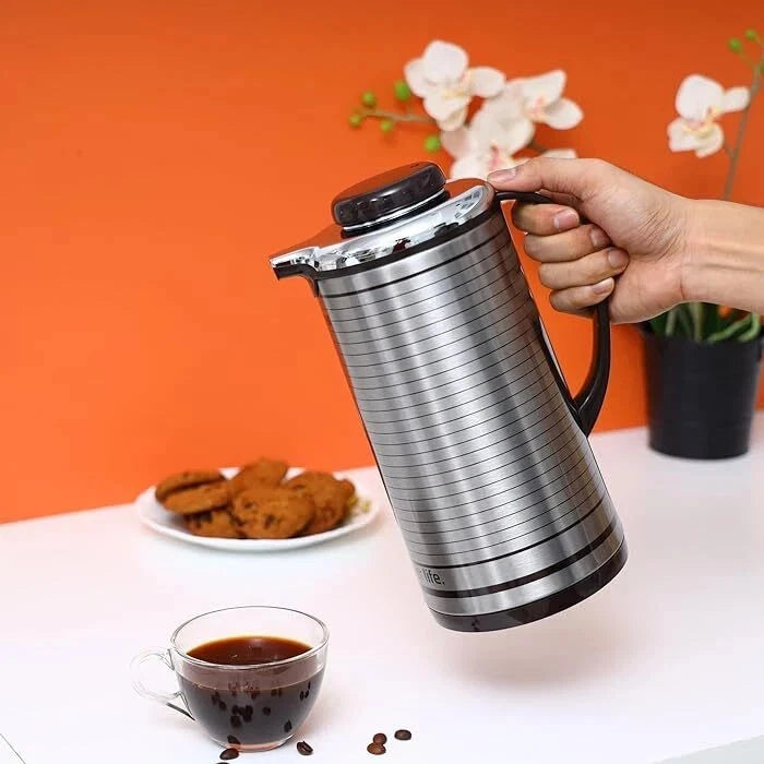 Person holding a silver thermal coffee press with cookies and a cup of coffee on a table.