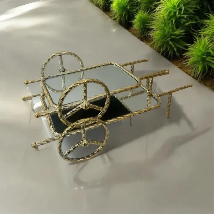 Metallic wheelbarrow sculpture on a reflective surface with greenery in the background